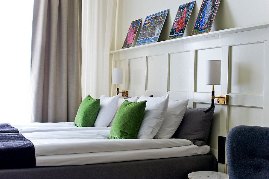 Fotografi av en fint bäddad säng med sänglampor på varsin sida samt fyra tavlor på väggen ovanför sängen.