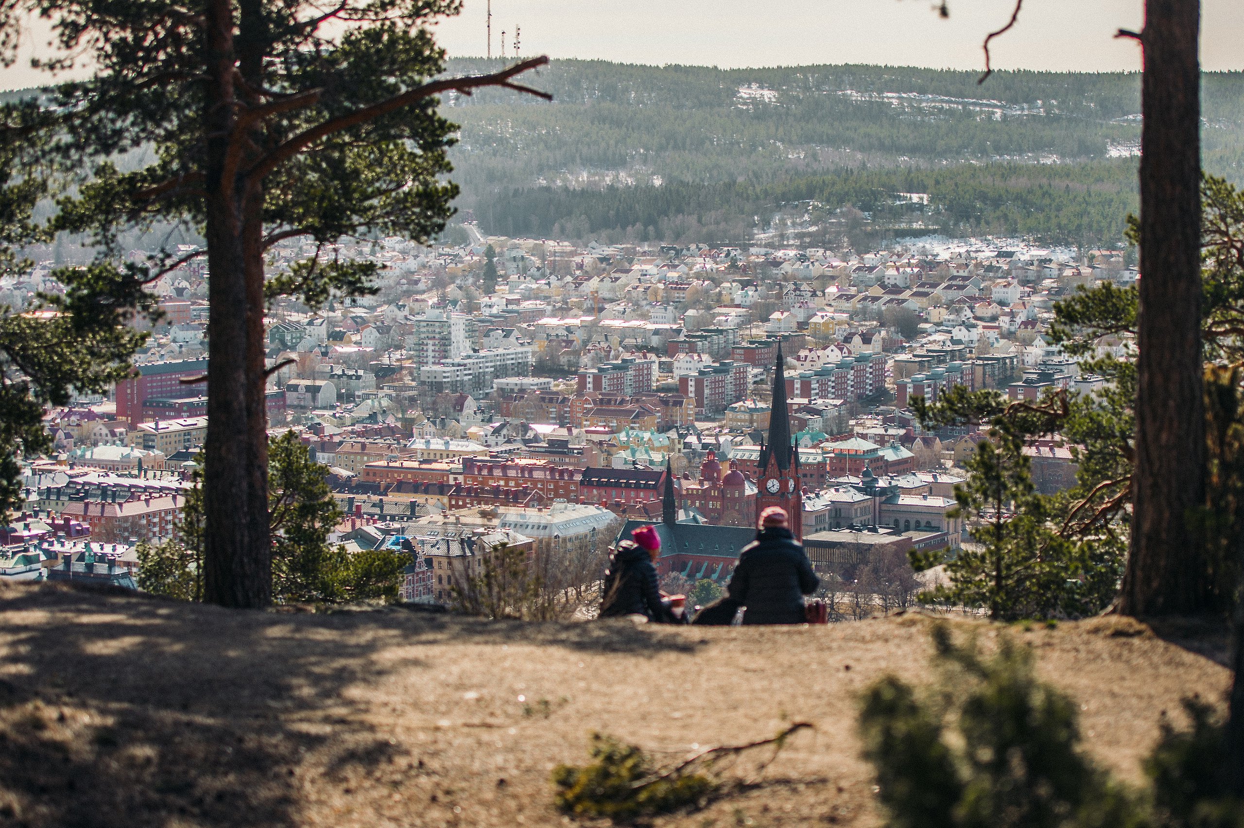 Fotografi av en vy över Sundsvalls stad. I förgrunden av bilden ser man ryggen av två personer som sitter och tittar ner på staden. 