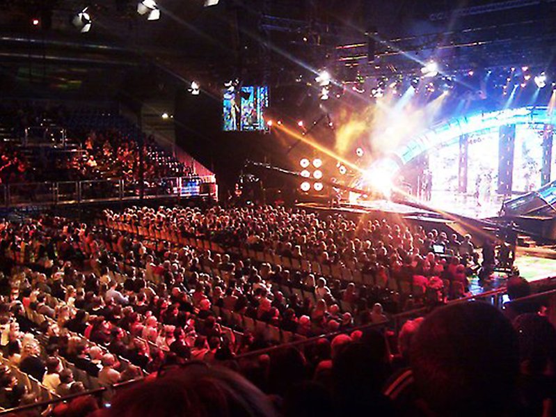 Fotografiet är fotat ovanifrån i en stor arena och man ser flera personer sitta och kolla upp på en scen som är upplyst. 