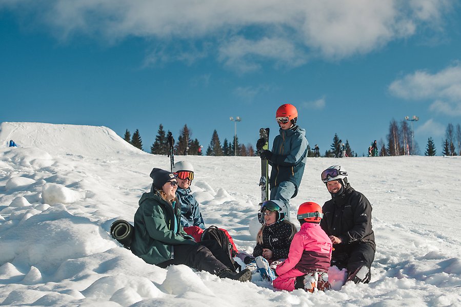 Fotografi av fem personer som sitter i slalombacken och en person står bredvid. Alla personer har skidkläder på sig och det är fint väder. 