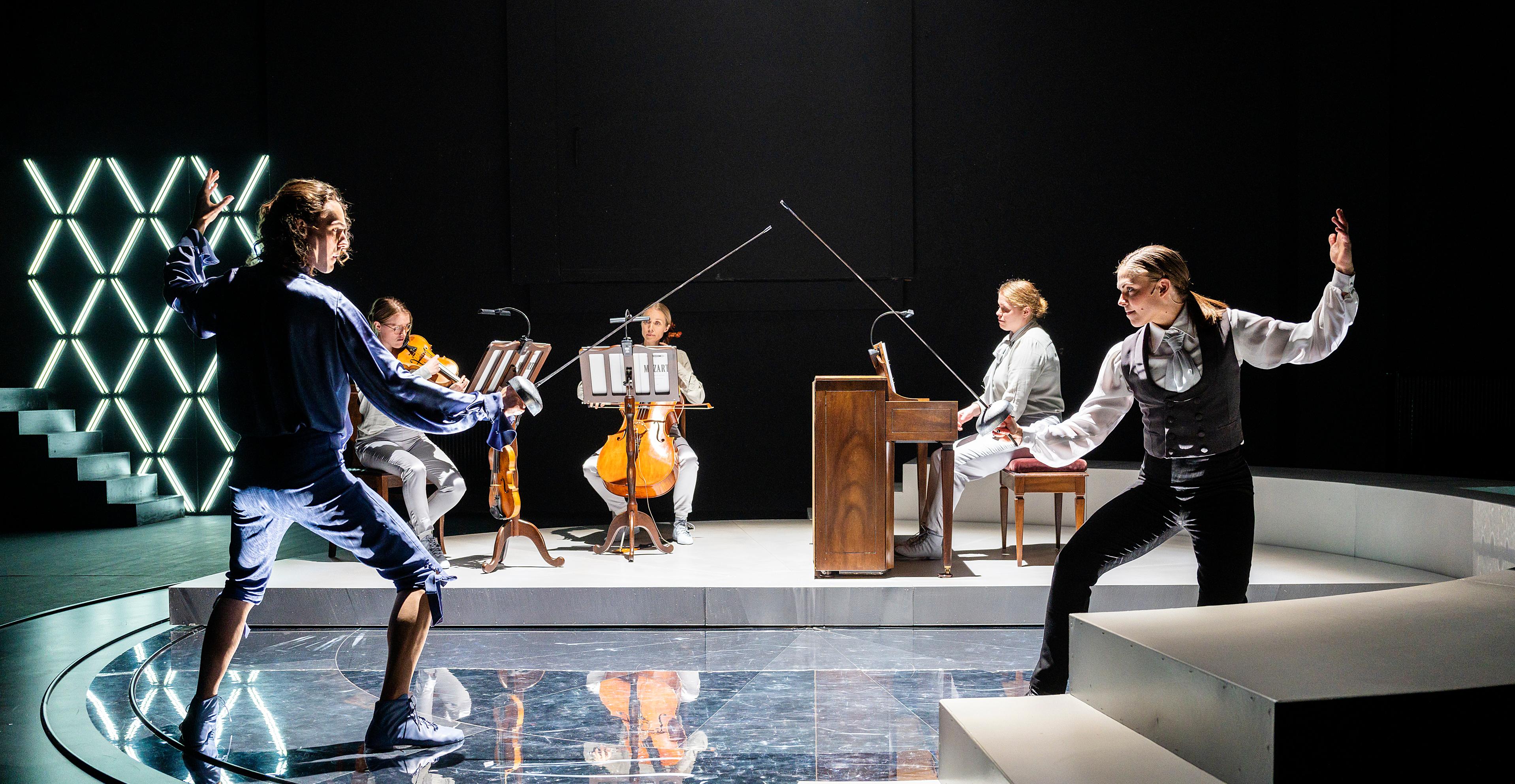 Ett foto. Två personer fäktas med silvriga värjor. De står på en scen med tre musiker i bakgrunden. Musikerna spelar fiol, cello och piano. 