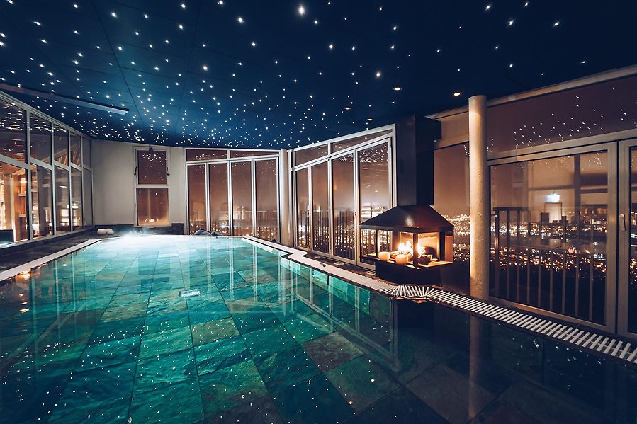Fotografi av en pool inomhus. Taket är fylld med små lampor som ser ut som stjärnor. Till höger om bilden bredvid poolen är en kamin.