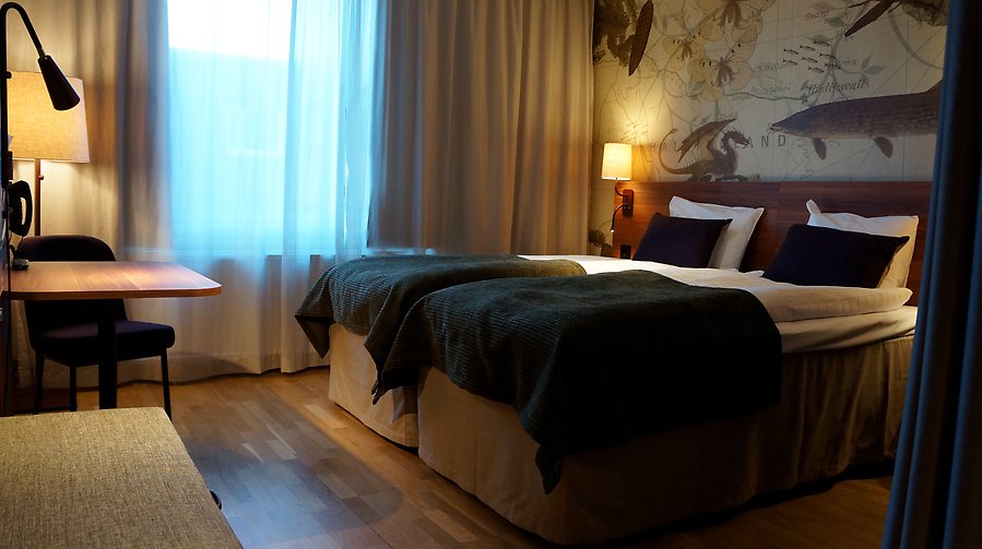 Fotografi av ett hotellrum med en dubbelsäng till höger i bild och ett skrivbord till vänster i bild.
