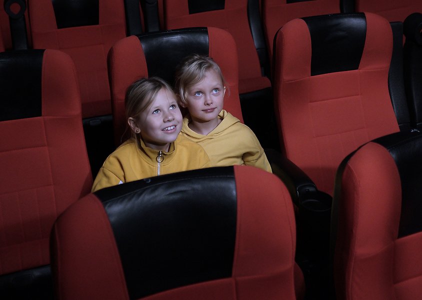 Två flickor sitter i en biostol i en tom biosalong. Sätena är röda och har svart skinn där man har huvudet. Flickorna är iklädda gula tröjor och de sitter och ler mot den stora bioduken.