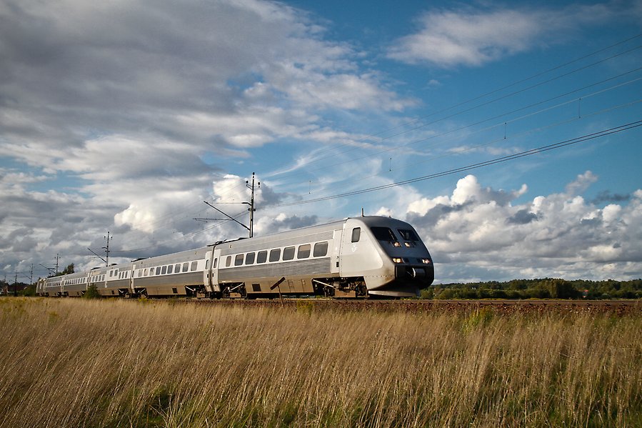 Ett silvrigt tåg kör i full fart på en järnväg som ligger längs med en äng.
