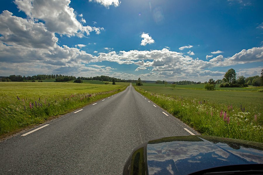 Ett foto på en lång och rak asfaltsväg med vita linjer. På vägen kör en svart bil och på båda sidor av vägen sträcker sig en stor grön lägda.