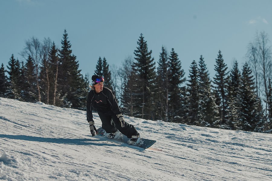 En man åker snowboard i en brant slalombacke. Pisten är snöigt vit och mannen är iklädd svarta kläder och har ett par stora glasögon på huvudet. 